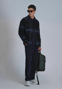 Veste en flanelle à carreaux noir et bleu, haut marine, pantalon marine, baskets grises, tenant un sac à dos vert avec des accents noirs, lunettes de soleil.