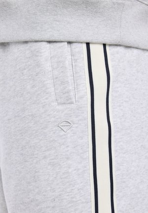 Pantalon de jogging gris clair avec poche latérale, bande blanche verticale bordée de fines lignes noires et petit logo géométrique brodé sous la poche.