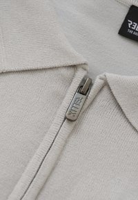 Lys grå strikpullover med en lynlåsåbning i kraven. Har en sølvfarvet lynlås med "RITTLER" indgraveret. Glat tekstur med ribbede kanter.