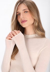 Lichtbeige blouse met lange mouwen en een hoge halslijn, met verlengde mouwen die de handen bedekken en een zachte, gladde textuur.
