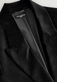 Černý sametový blazer s klopovým límcem, elegantními klopami a hladkou saténovou podšívkou. Jemná textura s minimálními švy.
