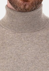 Homme barbu portant un pull à col roulé en tricot beige, montrant des détails en gros plan du tissu et du col.