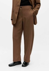 Rudos pritaikyti pločio kojų kelnes su aukštu liemeniu, derinamos su atitinkančiu švarku ir juodais odiniais loafers, dėvimi stovi. 