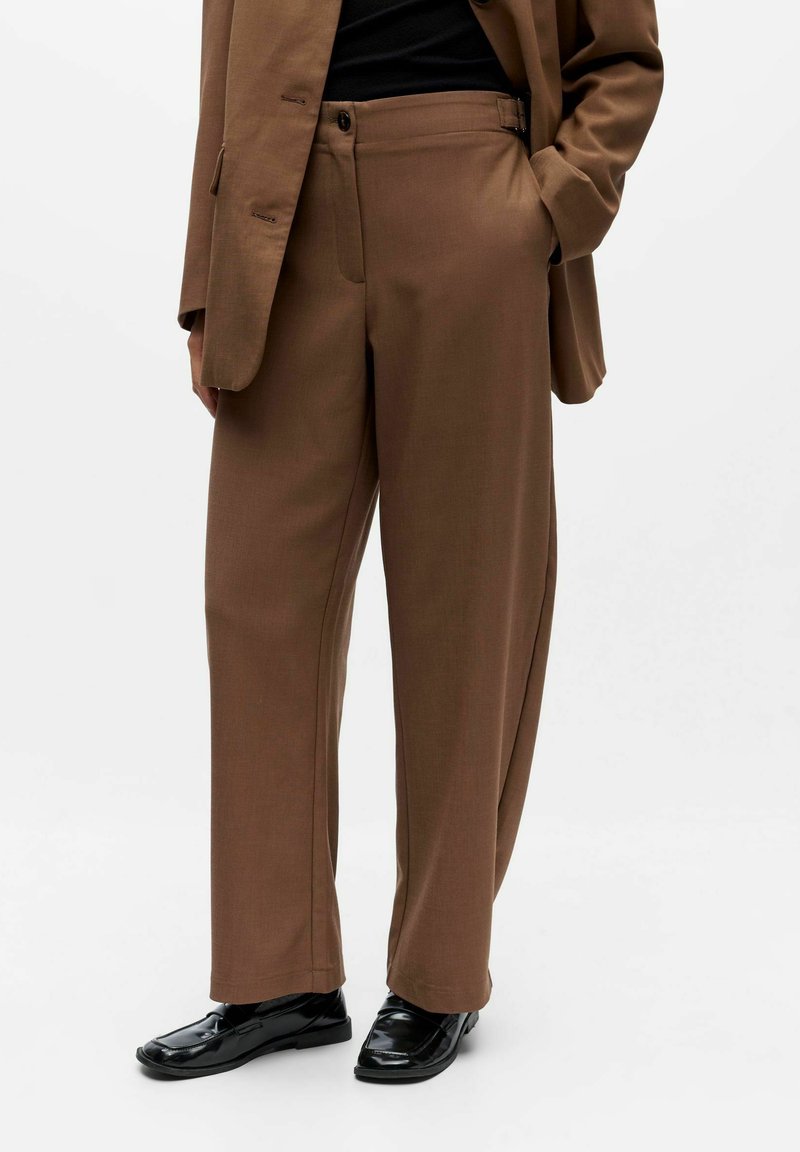 Rudos pritaikyti pločio kojų kelnes su aukštu liemeniu, derinamos su atitinkančiu švarku ir juodais odiniais loafers, dėvimi stovi. 