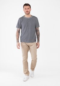Hombre con tatuajes en los brazos avanza luciendo una camiseta de rayas blancas y negras, pantalones beiges y zapatillas blancas sobre un fondo blanco.