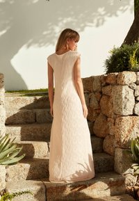 Robe nude longue et texturée avec des manches courtes, présentant des rayures verticales. Le mannequin se tient sur des marches en pierre avec de la verdure et un mur blanc en arrière-plan.