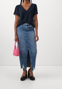 Blouse bleu marine à col en V avec manches courtes à volants, associée à une jupe en denim taille haute avec une fente à l'avant et un ourlet effiloché ; tenant un sac rose.
