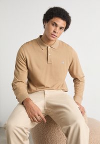 Polo de manga larga beige con cuello y tapeta de botones, que presenta un pequeño logo en el pecho. Acompañado de pantalones beige claros.