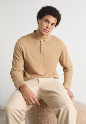 Beige långärmad polo med krage och knapprad, med en liten logotyp på bröstet.  Matchas med ljusbeige byxor.