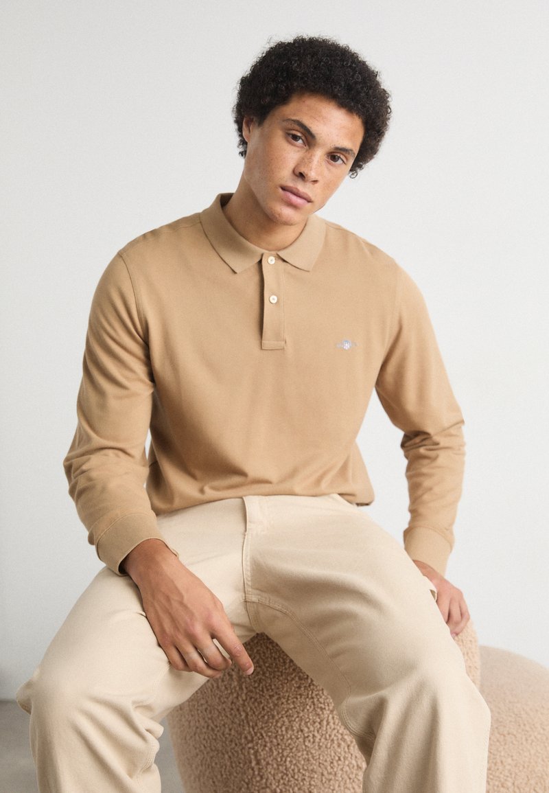 Polo de manga larga beige con cuello y tapeta de botones, que presenta un pequeño logo en el pecho. Acompañado de pantalones beige claros.