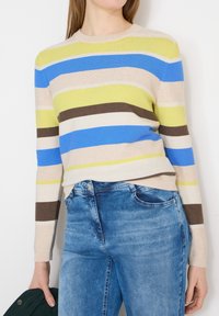 Femme portant un pull rayé à manches longues bleu, jaune, marron et beige, associé à un jean bleu, tenant une veste vert foncé.