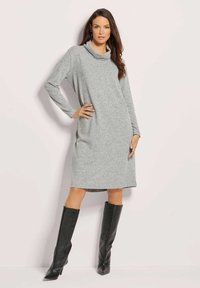 Graues Kleid mit Kapuzenkragen, langen Ärmeln und seitlichen Taschen. Aus glattem Strickstoff und in mittellanger Passform. Kombiniert mit schwarzen kniehohen Stiefeln.