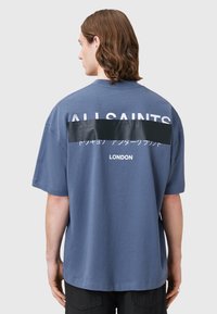 Blå bomulds T-shirt med korte ærmer, der har stor hvid tekst "ALL SAINTS" og en sort vandret stribe på ryggen.
