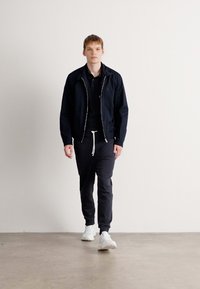 Ung mand går indendørs iført en sort jakke, sort polo, sorte joggers med hvid snøre og hvide sneakers foran en ensfarvet hvid væg.