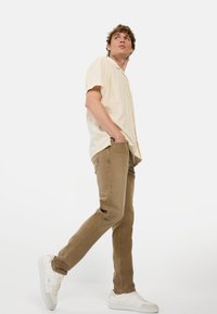 Lichtbeige korte mouwen shirt, tan slim-fit broek en witte sneakers. Het shirt heeft een knoopsluiting; de broek heeft subtiele stiksels.
