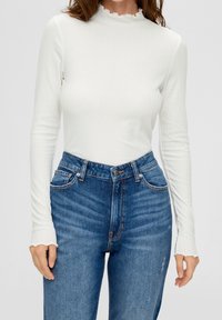 Bílý žebrovaný top s dlouhými rukávy a vysokým rýhovaným výstřihem, v kombinaci s modrými džíny s vysokým pasem a tradičním pěti-pocket designem.