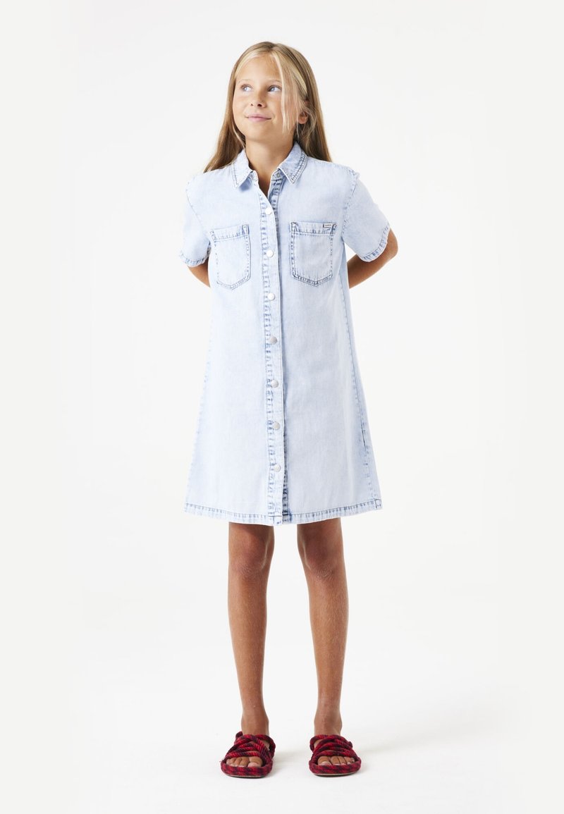 Hellblaues Denim-Hemdkleid mit kurzen Ärmeln, vorderem Knopfverschluss, zwei Brusttaschen und geradem Saum. Getragen mit roten Sandalen.