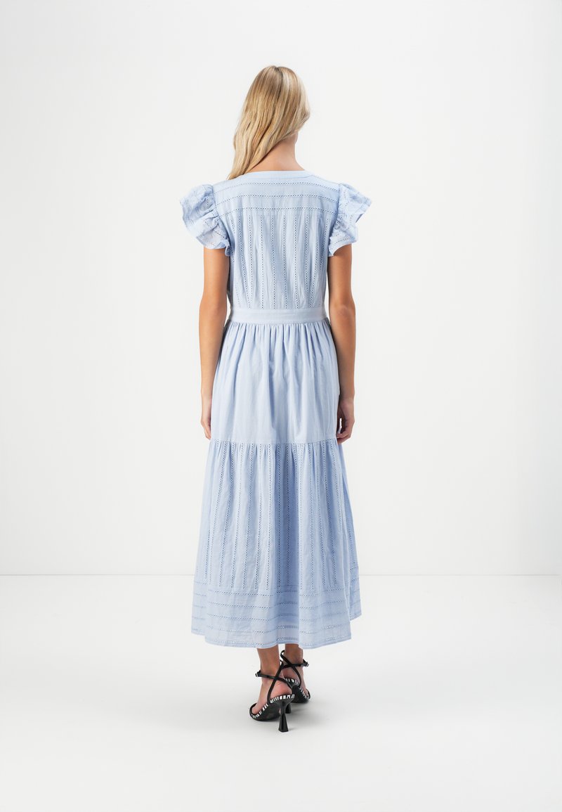 Kobieta z długimi blond włosami, ubrana w jasnoniebieską sukienkę midi z falbaniastymi rękawami i czarnymi sandałami na wysokim obcasie z paskami, stojąca na białym tle.