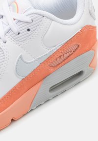 Vita läder sneakers med orange mocka detaljer, grå Nike-logga, stor luftkudde och gummisula. Har en texturerad snörningsyta och sömmar.