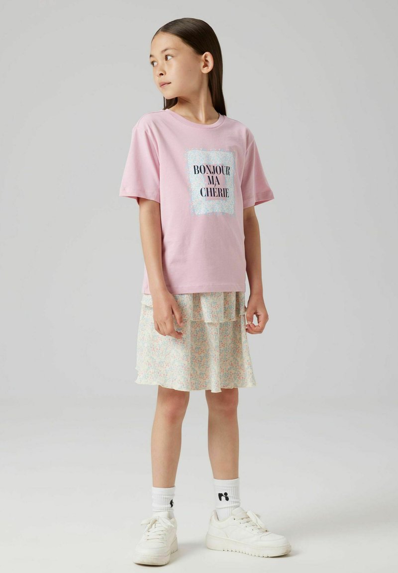 T-shirt di cotone rosa chiaro con "BONJOUR MA CHERIE" stampato in nero su uno sfondo glitterato, abbinato a una gonna a strati con motivo floreale.