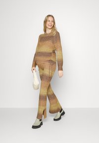 Sweater de malha e calças combinando em gradiente de castanho e amarelo. O suéter tem franzidos laterais; as calças apresentam aberturas e perna estilizada. Bolsa bege e botas.