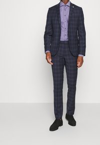 Námořnicky modrý kostkovaný oblek s úzkým střihem, doplněný světle fialovou košilí na knoflíky a černými botami. Látka obleku má hladkou texturu a strukturovaný design.