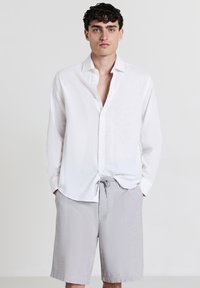 Jeune homme avec des cheveux bouclés et foncés portant une chemise blanche à manches longues ouverte et un short gris clair à cordon de serrage, se tenant devant un fond uni.