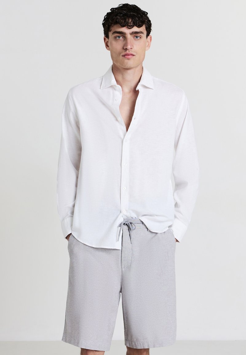 Jeune homme avec des cheveux bouclés et foncés portant une chemise blanche à manches longues ouverte et un short gris clair à cordon de serrage, se tenant devant un fond uni.