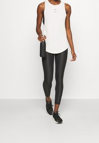 Bílý sporty bezramenný top, černé legíny se vzorem a černé tenisky. Modelka držící černou láhev na vodu s elegantním, minimalistickým designem.