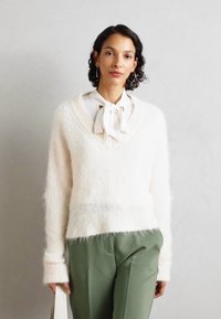 Pulover pufos alb cu decolteu adânc în V, asortat cu o bluză lejeră dotată cu un gât legat și pantaloni verzi măslinii. Design simplu, elegant.