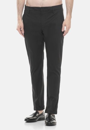 Uomo che indossa pantaloni neri a taglio aderente e scarpe slip-on in pelle nera, in piedi contro uno sfondo chiaro e semplice.