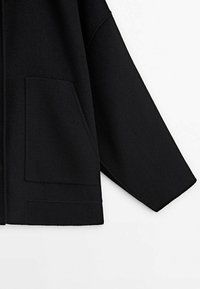 Manteau noir en tissu épais et doux, avec une coupe décontractée, des manches larges et deux poches avant, présentant un design épuré et minimaliste.