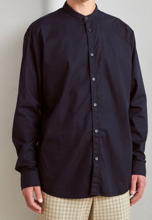 Homme portant une chemise à col mandarin bleu marine foncé avec un pantalon à carreaux beige, se tenant contre un mur clair.