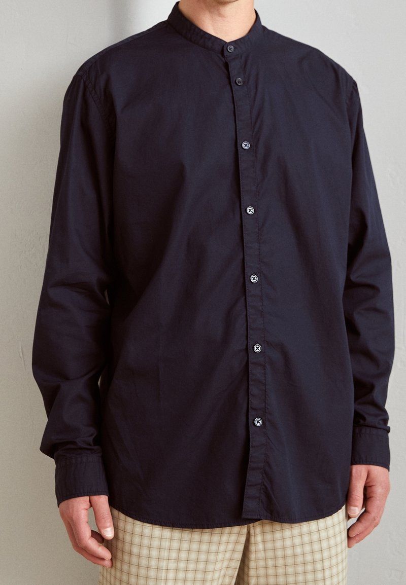 Muž v tmavě modré košili s mandarin límečkem a béžových károvaných kalhotách stojí před hladkou světlou stěnou.