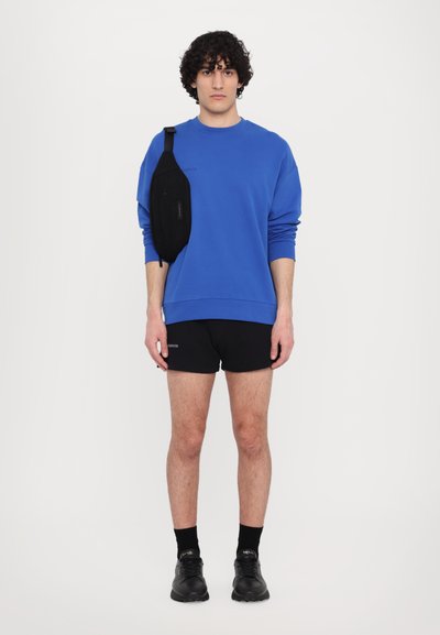 Felpa blu, pantaloni corti neri, marsupio nero indossato a tracolla, sneakers nere, modello in piedi su uno sfondo bianco semplice.
