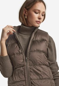 Braune Steppweste mit Quiltmuster, hohem Kragen und Reißverschluss, getragen über einem passenden Rollkragenpullover. Glattes, glänzendes Material.
