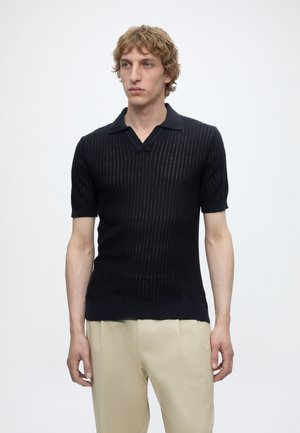 Νέος άνδρας με σγουρά ξανθά μαλλιά, που φοράει μαύρο ριπ κοντομάνικο πόλο και μπεζ παντελόνια, στέκεται μπροστά σε λευκό φόντο.