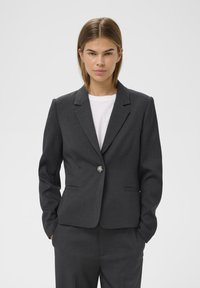Szürke, szabott blézer egy gombos záródással, kivágott hajtókákkal és két elülső zsebbel, texturált anyagból. Fehér ing felett viselve.