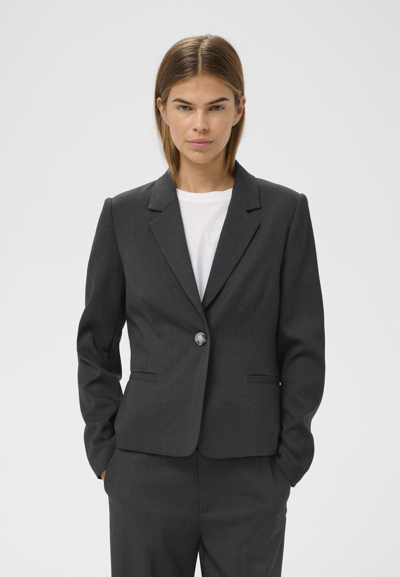 Szürke, szabott blézer egy gombos záródással, kivágott hajtókákkal és két elülső zsebbel, texturált anyagból. Fehér ing felett viselve.