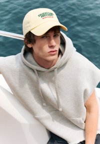 Lys grå ærmeløs hoodie med en lomme, parret med en gul kasket med grøn og rød broderet tekst, mod en blå vandbaggrund.