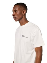 Mann mit kurzen Haaren, der ein weißes T-Shirt mit dem Text "HALO 196819721981" auf der Brust trägt, nach links blickend vor einem einfarbigen Hintergrund.
