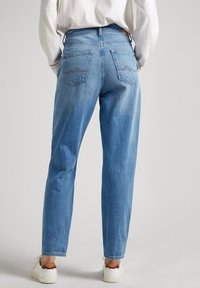 Person klädd i ljusblå raka jeans och vita sneakers, stående med händerna i fickorna mot en enkel bakgrund.