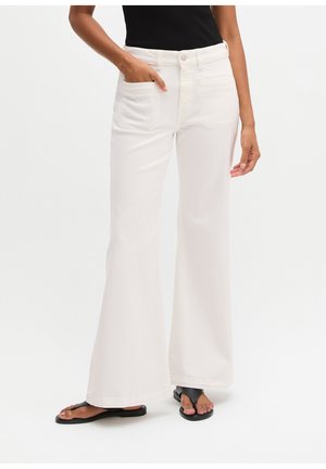 Kobieta nosząca białe spodnie z wysokim stanem i szerokimi nogawkami z przednimi kieszeniami, w połączeniu z czarnymi klapkami i czarnym topem bez rękawów.