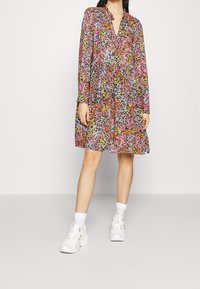 Floral gedrukte jurk met lange mouwen, een V-hals en een uitlopende silhouet. Kleuren zijn roze, oranje, blauw en zwart. G gedragen met witte sneakers.