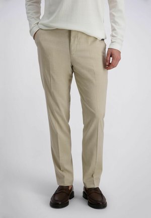 Mann trägt beige, schmal geschnittene Hose, weißes langärmeliges Hemd und dunkelbraune Leder-Loafer, steht mit einer Hand in der Tasche.