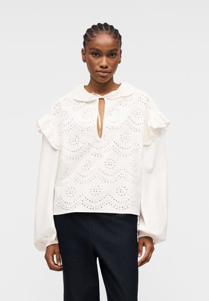 Frau mit geflochtenem Haar, die eine weiße Lochstickerei-Bluse mit gerafften Schultern und langen, puffigen Ärmeln trägt, kombiniert mit dunkler Hose.