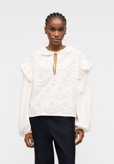 Donna con capelli intrecciati che indossa una blusa bianca di pizzo sangallo con spalle arricciate e maniche lunghe a sbuffo, abbinata a pantaloni scuri.