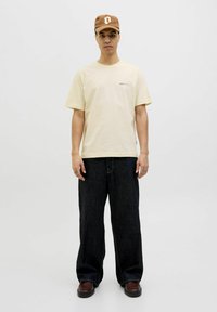 Beige cotton t-shirt with short sleeves, paired with dark wide-leg denim pants. Brown baseball cap featuring a white logo. Brown shoes.