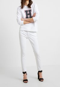 Sudadera blanca con un logo "H" en negro y rojo, combinada con jeans skinny blancos y sandalias de tacón negro. Textura de tela suave.