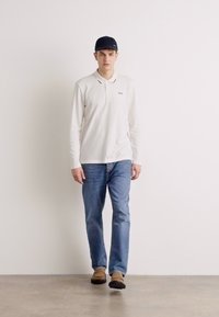 Bílá polo košile s dlouhými rukávy, modré džíny, hnědé boty a tmavý klobouk. Jednoduchý design s logem na košili, stojící na neutrálním pozadí.